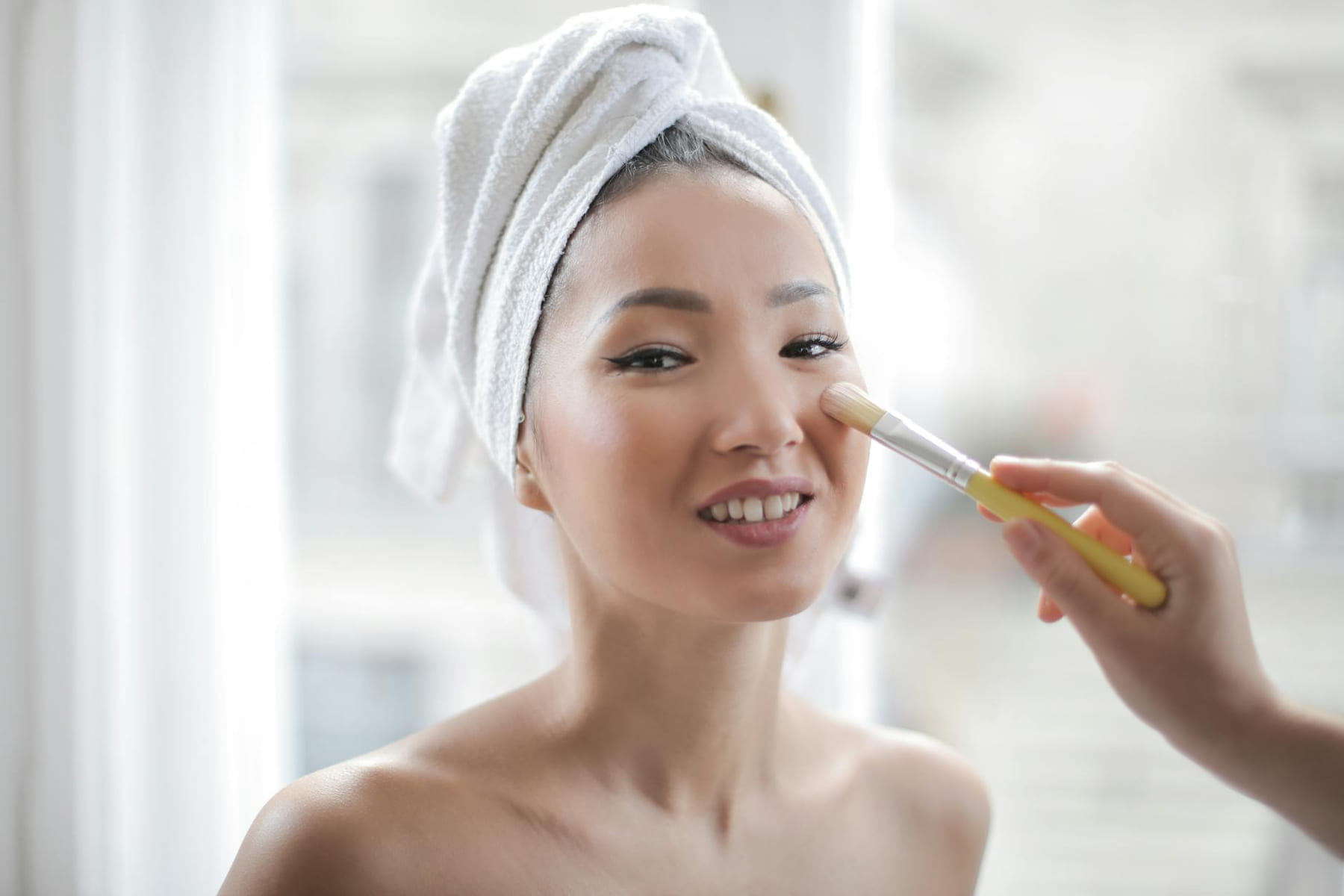 Smiling woman with a towel on her head enjoying a makeup session indoors, highlighting natural beauty and healthy skin. on Purely Polished