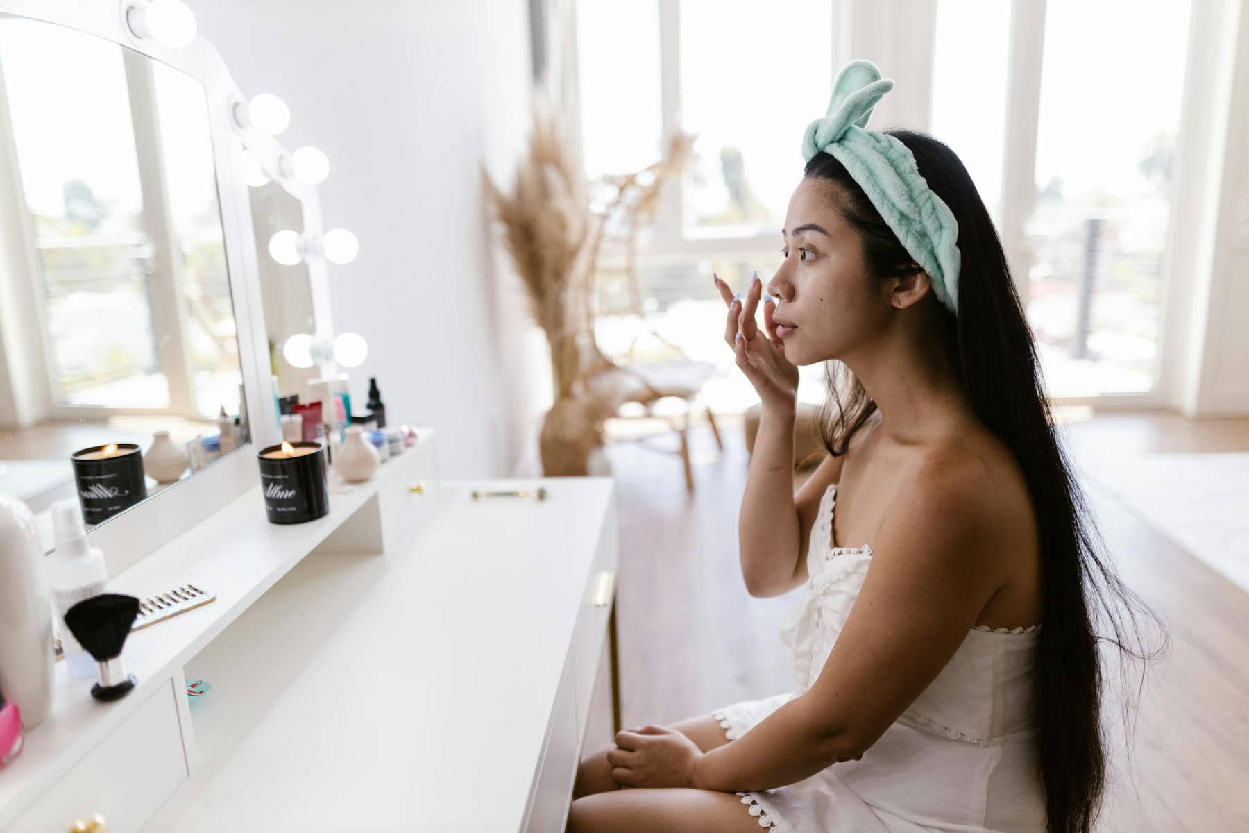 A woman applying makeup at a vanity mirror with a headband, candles, and beauty products. on Purely Polished