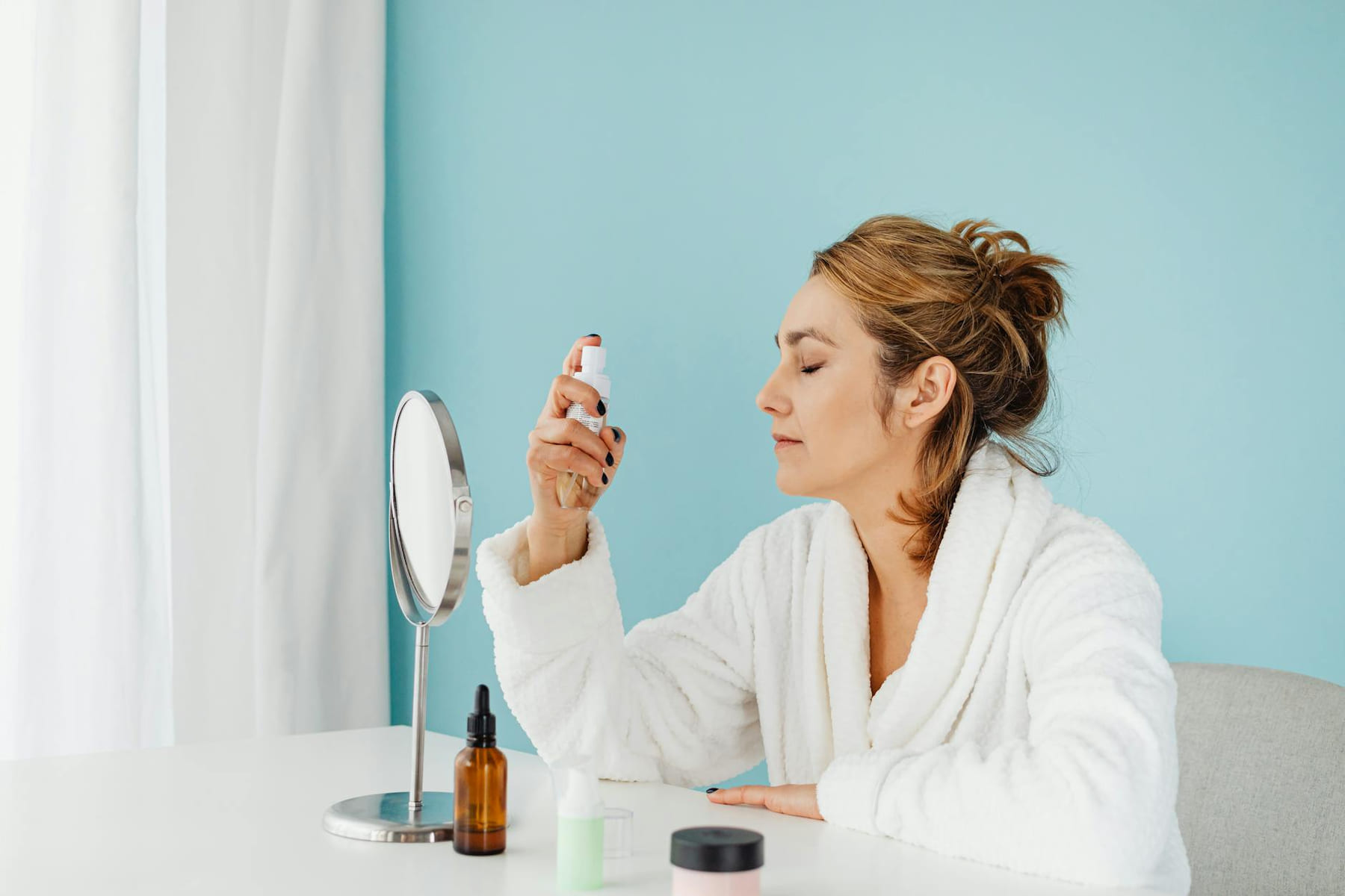 Woman in a bathrobe using a skincare spray in front of a mirror indoors. on Purely Polished