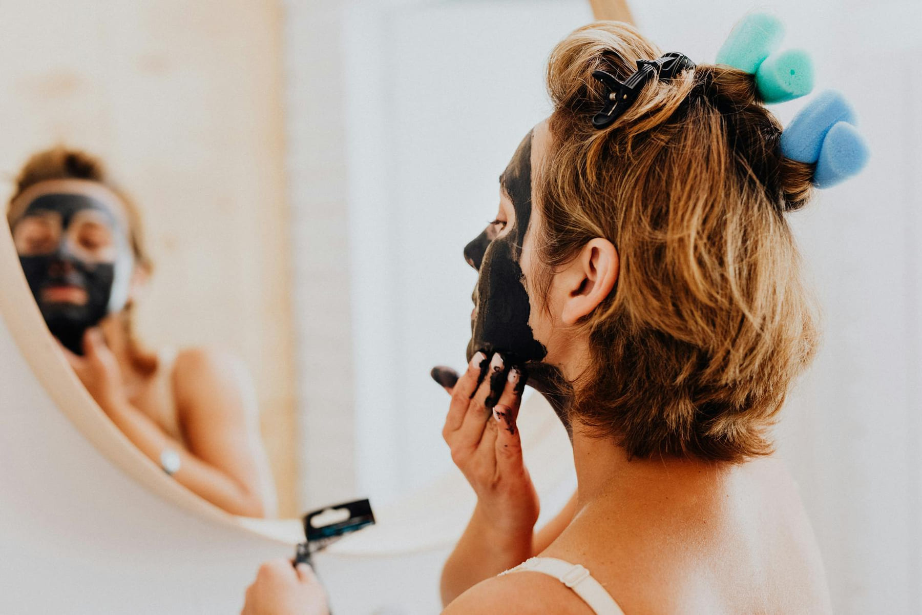 A woman is applying a black facial mask in front of a mirror, focusing on skincare and beauty routines. on Purely Polished