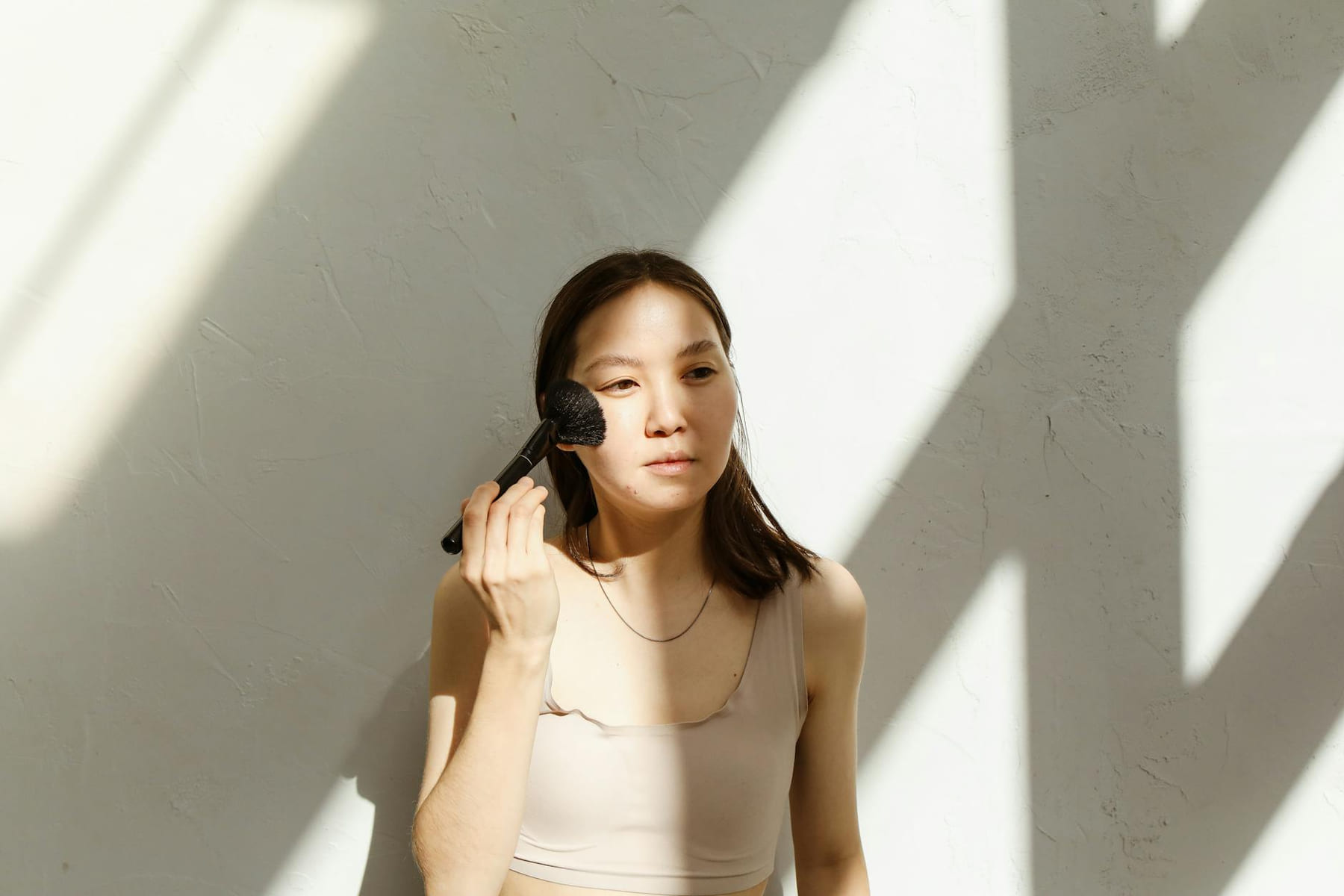 Asian woman applying makeup with brush in natural sunlight indoors. on Purely Polished