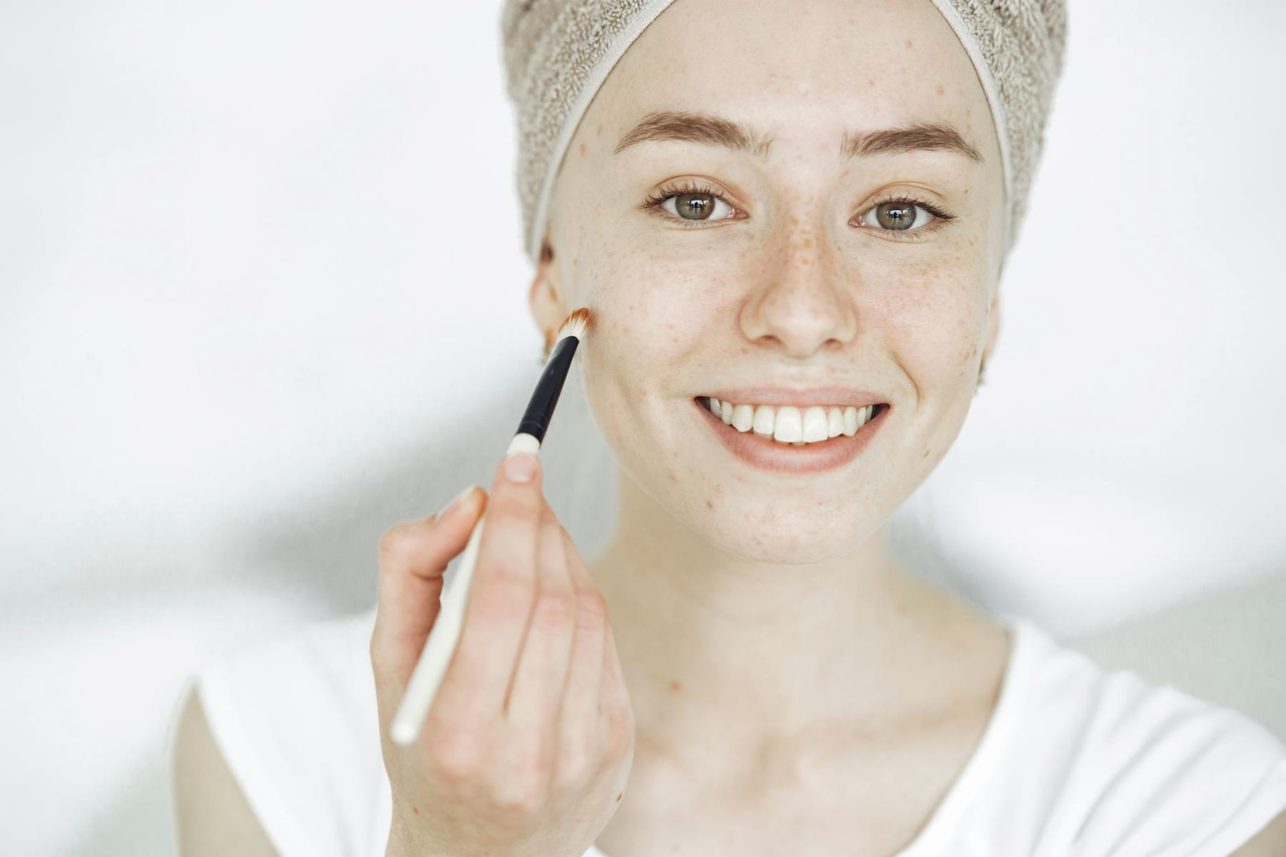 Close-up of a smiling woman applying makeup with a brush, enhancing natural beauty. on Purely Polished