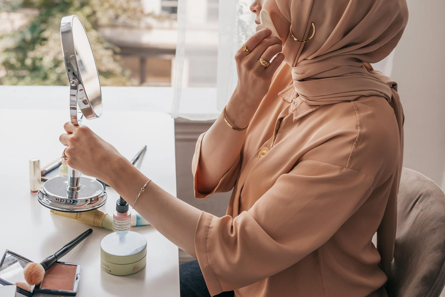 A woman in a hijab applies makeup at a bright indoor setting by a window. on Purely Polished