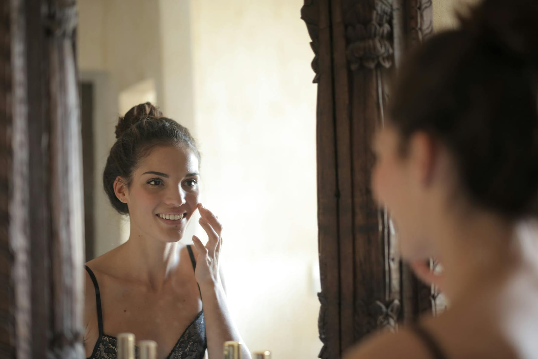 Young woman applying skincare and smiling at her reflection in a rustic mirror. on Purely Polished