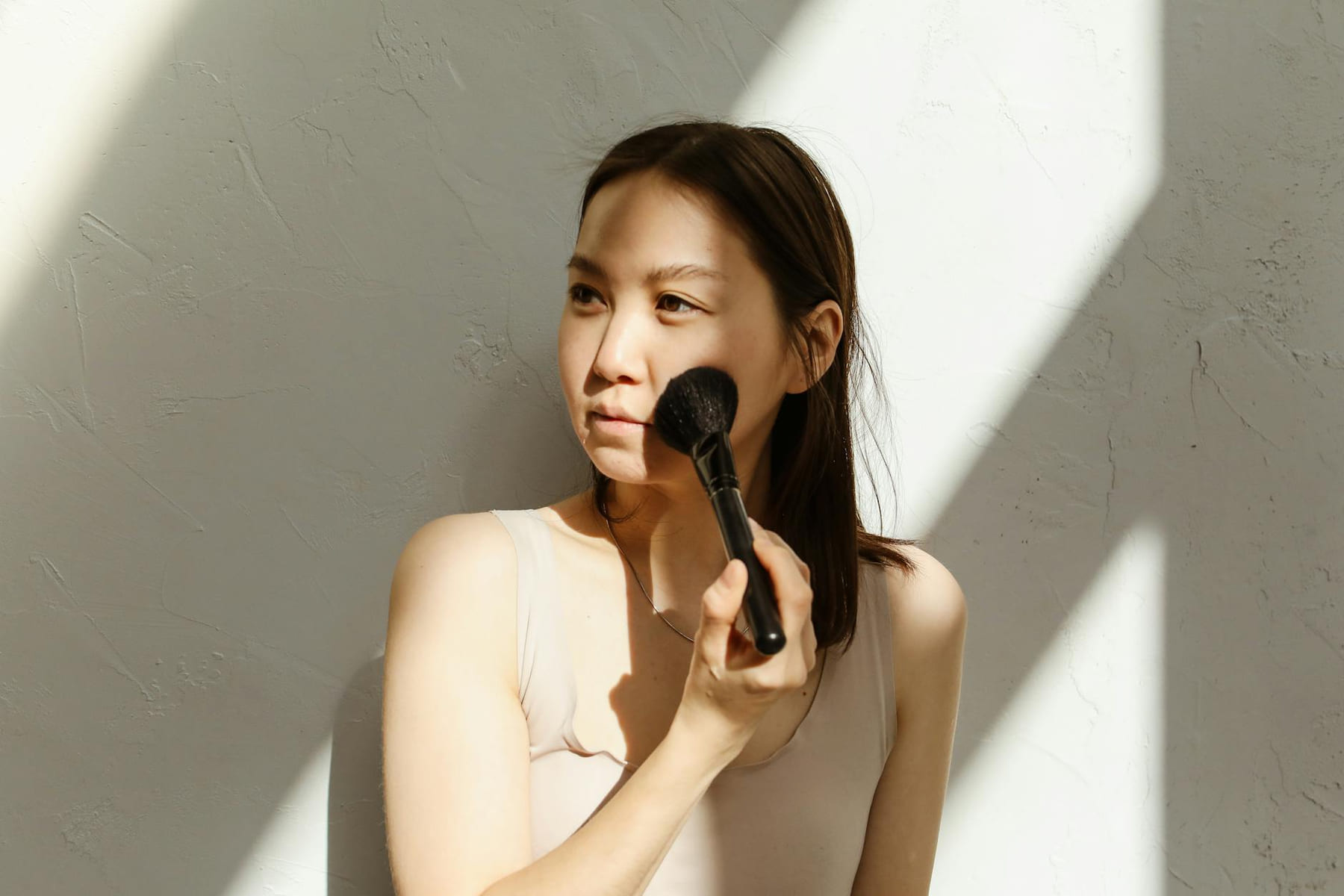 A young Asian woman applying makeup with a brush in natural lighting. on Purely Polished