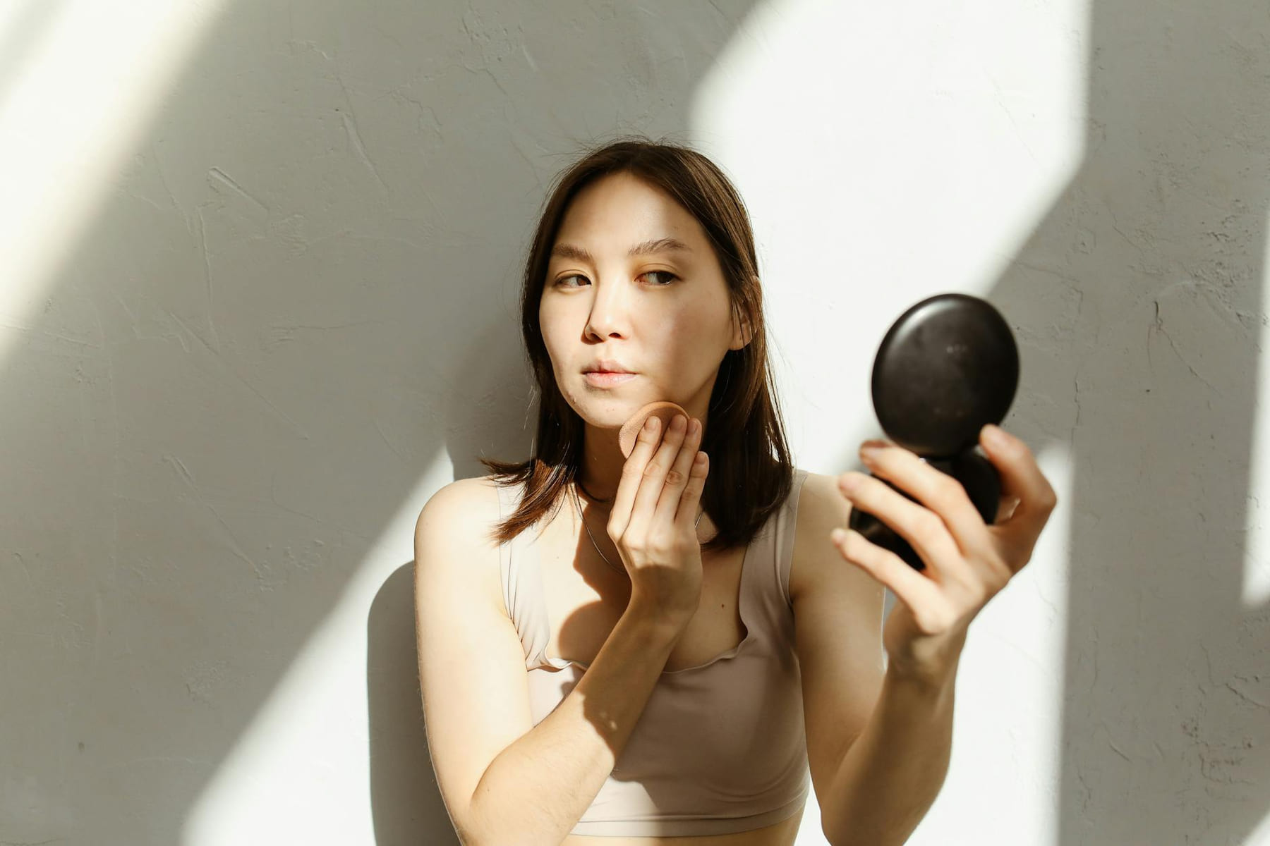 A young woman applying makeup, illuminated by natural light streaming through a window. on Purely Polished