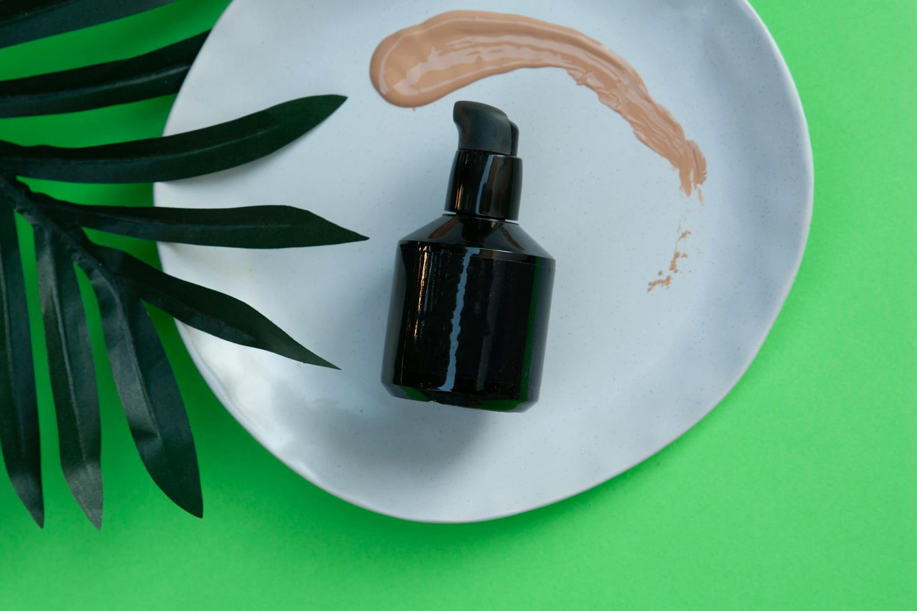 Black skincare pump bottle on a white plate with green leaf and makeup smear. on Purely Polished