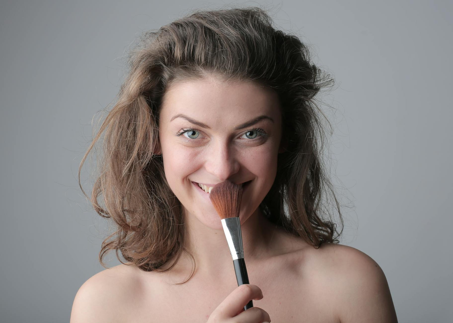 Close-up of a smiling woman holding a makeup brush against gray background. on Purely Polished